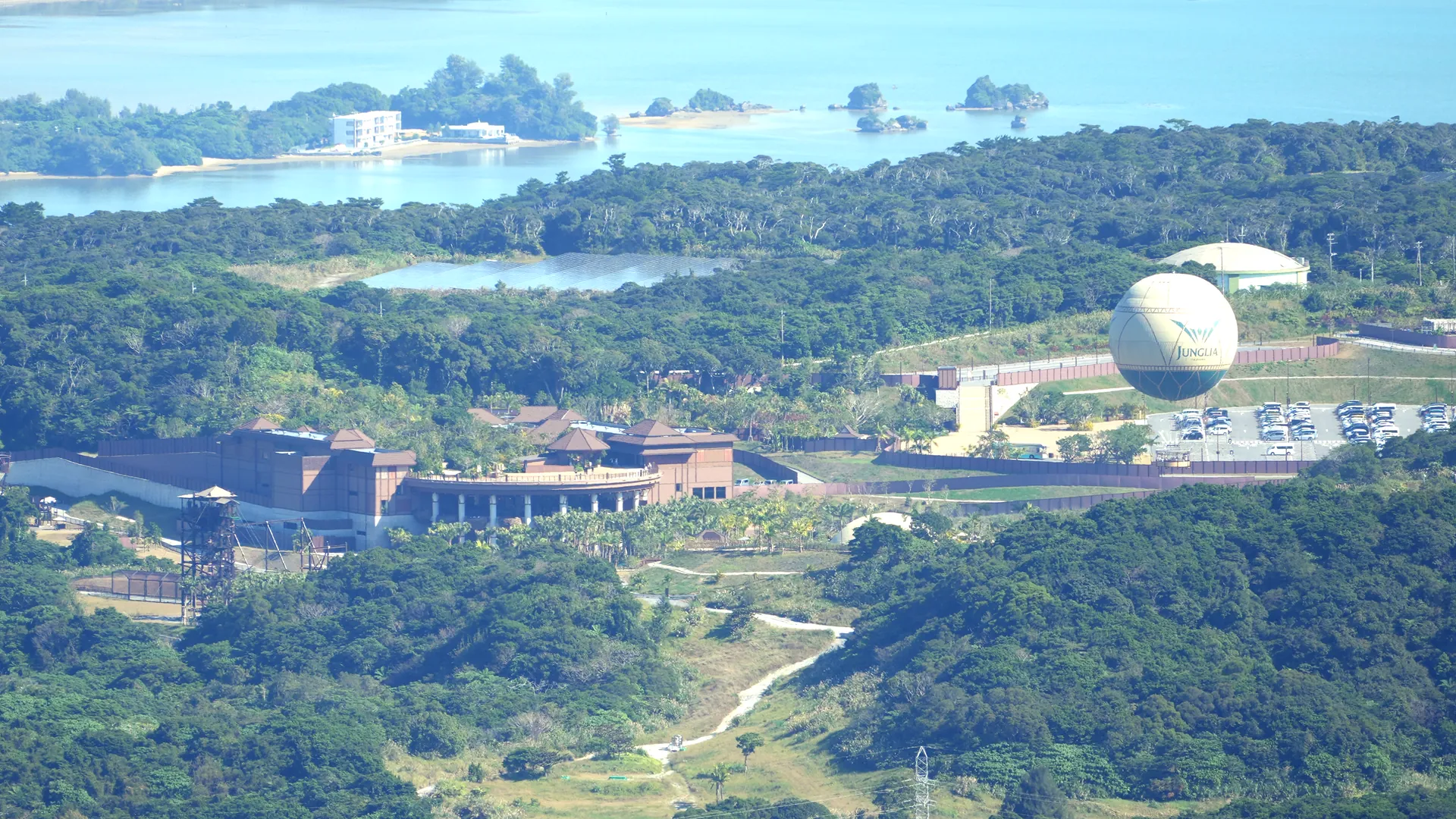 The park seen from Mount Yae: its entrance buildings and tethered balloon set in the Yanbaru forest, with a bay and small islands behind