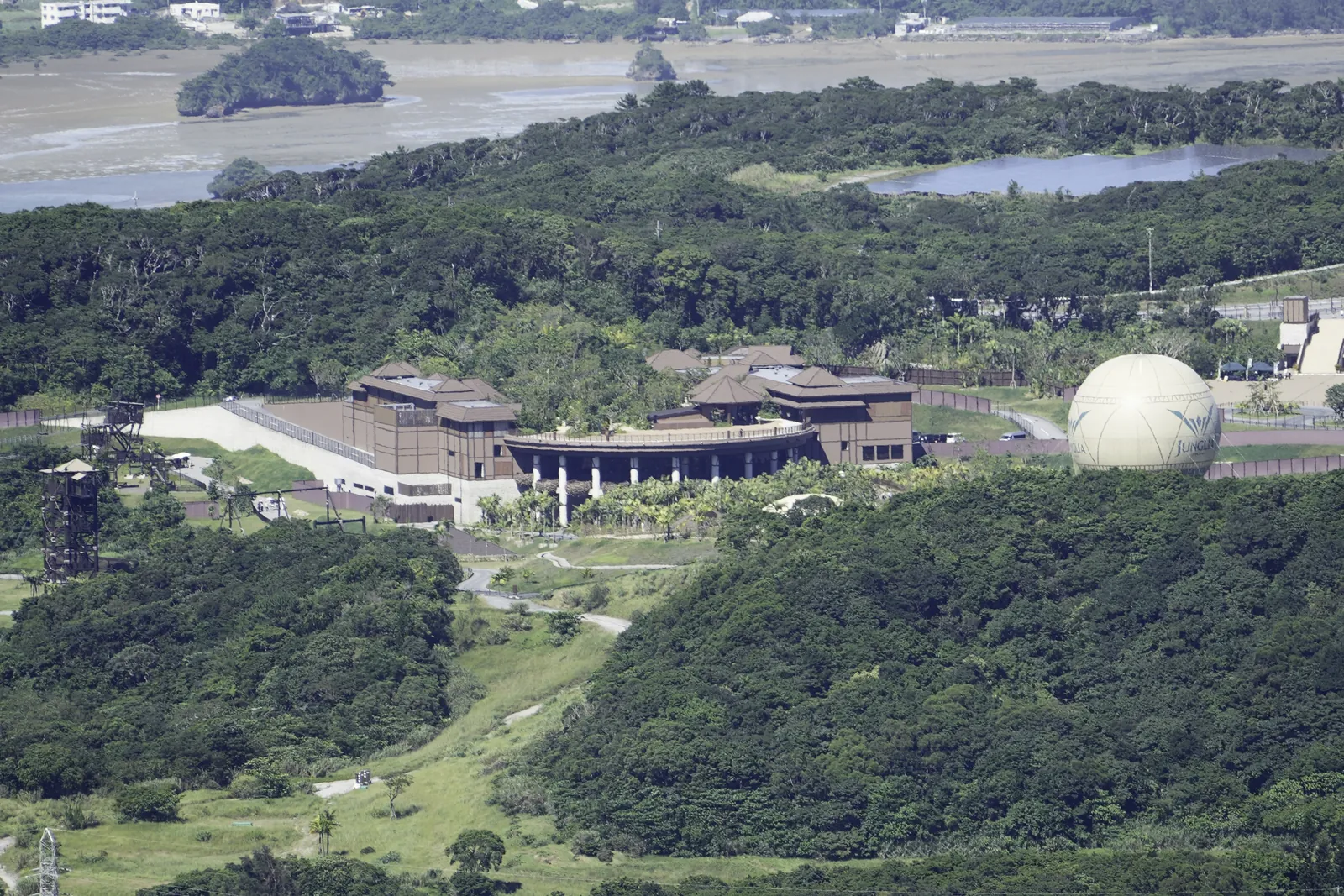 Parkens brune indgangsbygninger og en stor, lys ballon blandt tæt grøn skov, med en sø og en tidevandsbugt bagved, set fra en bakketop i det nordlige Okinawa